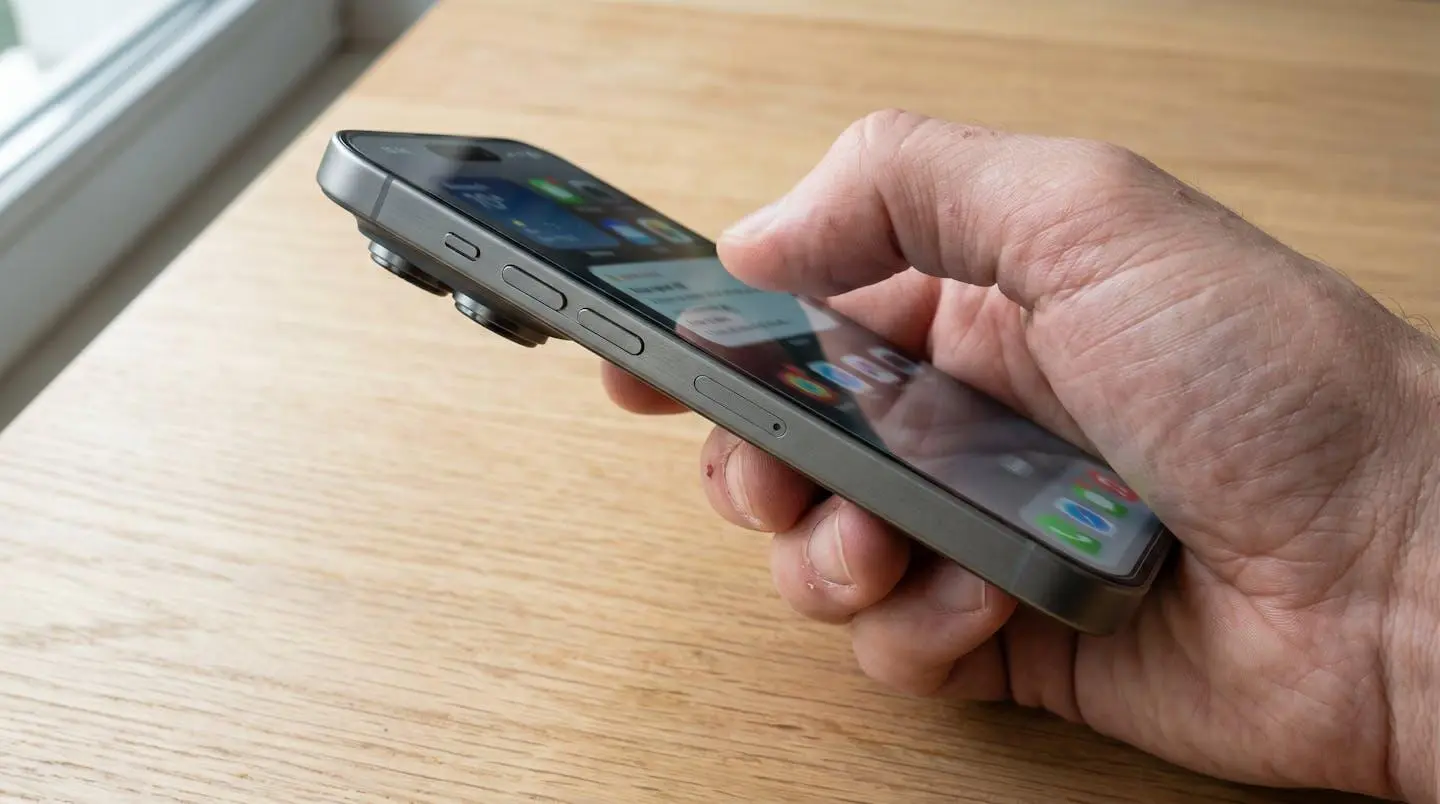Extreme close-up of a hand holding a modern smartphone with the screen partially visible and softly blurred, set against a clean contemporary surface with beautiful background bokeh